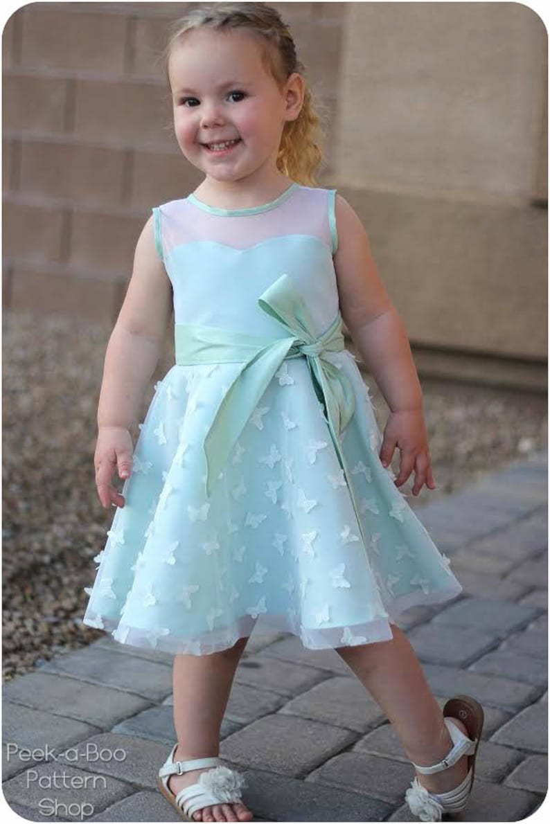 May include: A young girl wearing a light blue dress with a satin sash and butterfly embellishments. The dress has a sheer bodice and a full skirt. The girl is smiling and looking at the camera.