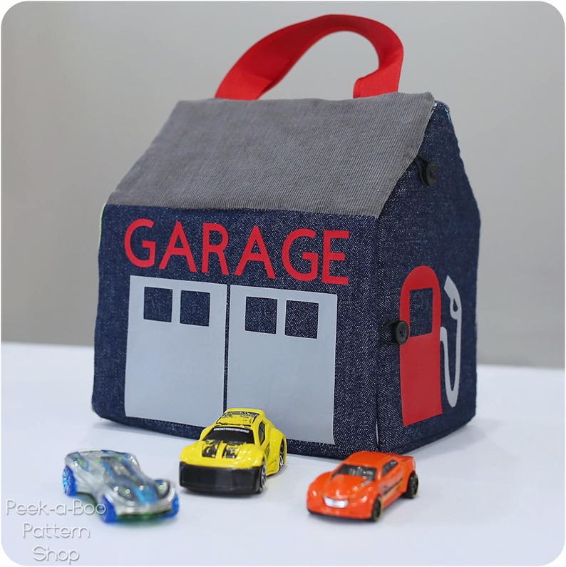 Peut inclure: Un garage en jean bleu avec une poign&eacute;e rouge et le mot "GARAGE" en lettres rouges. Le garage a deux portes grises et une pompe &agrave; essence rouge sur le c&ocirc;t&eacute;. Il y a trois voitures miniatures devant le garage, une jaune, une bleue et une orange.