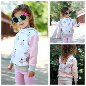 May include: A pink and white hooded sweatshirt with a penguin print. The sweatshirt has a kangaroo pocket and a contrasting pink sleeve. The model is wearing pink leggings and sunglasses.