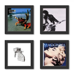 May include: Four square black frames displaying various music album covers, including The Prodigy, Bruce Springsteen, and Madonna. One frame is shown open with a hinge mechanism, revealing a circular cutout for a vinyl record inside the frame.