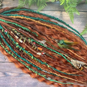 Puede incluir: Conjunto de rastas de color marrón rojizo con detalles en verde azulado y marrón. Las rastas están adornadas con elementos decorativos como hojas, cuentas y plumas, creando una estética bohemia. Sobre un fondo de madera.