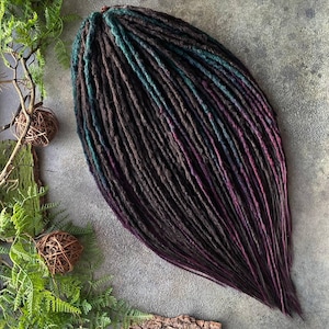 May include: A collection of dreadlocks in brown, teal, and purple hues. The dreads are arranged in a cascading style, with the colours blending from top to bottom. The background is a neutral grey, with natural elements such as greenery and a pine cone.