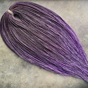 May include: A bundle of purple and dark brown synthetic dreadlocks, tied together with a light brown band. The dreadlocks have a gradient effect, transitioning from dark brown to vibrant purple. The hair is arranged on a gray surface.