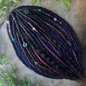 Op de afbeelding: Donkerblauwe en zwarte dreadlocks met paarse en blauwgroene strepen. De dreadlocks zijn versierd met kralen, knopen en zilveren bedels. De dreadlocks zijn gerangschikt op een neutrale ondergrond met groene varenbladeren.