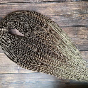 May include: A bundle of brown and blonde hair extensions, tied together at one end. The hair is arranged in a fan shape, with the colors blending from dark brown to light blonde. The extensions are lying on a wooden surface.
