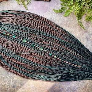 May include: A collection of brown and teal dreadlocks, adorned with a beaded strand featuring green and clear beads. The dreadlocks are arranged in a fan shape, with a gradient effect from brown to teal. The background is a textured, neutral surface.