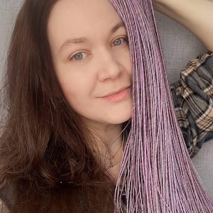 Puede incluir: Un conjunto de extensiones de cabello largas y finas que presentan una mezcla de fibras de color morado claro, lavanda y blanco. Los mechones están fuertemente trenzados en cordones individuales, creando una apariencia texturizada y tonal.