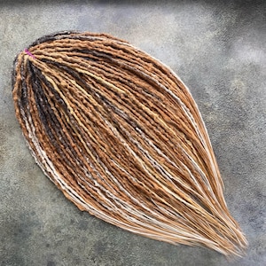 May include: A set of synthetic dreadlocks featuring a blend of dark brown, medium brown, and light blonde tones. The individual strands are textured and twisted, gathered at the top with a small pink elastic band, and taper into loose ends.