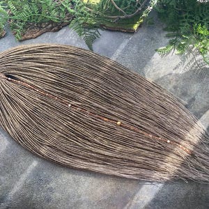 Puede incluir: Un conjunto de rastas marrones, adornadas con pequeñas cuentas, descansa sobre una superficie gris. Las rastas están dispuestas en forma de lágrima, con un aspecto natural y texturizado. El follaje verde enmarca la imagen.