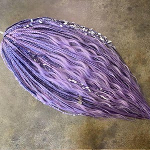 May include: A collection of lavender and purple synthetic hair braids. The braids are decorated with white accents and small silver-coloured embellishments. The hair changes from tightly braided to wavy at the ends.