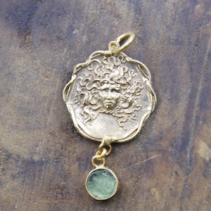 May include: A gold-coloured pendant depicting Medusa's face in relief, with detailed hair. A small, round, light green gemstone hangs below. The pendant has a textured, antique appearance, with a loop for a chain.