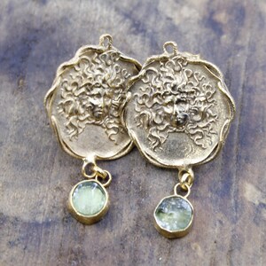 May include: A pair of gold-coloured earrings. Each earring features a detailed face design with curly hair and a small, round, light green stone pendant. The earrings are displayed on a wooden surface.