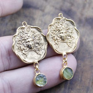 May include: A pair of gold-coloured earrings. Each earring features a detailed Medusa face design with flowing hair. A small, round, green gemstone hangs below each Medusa pendant, adding a pop of colour and contrast to the design.