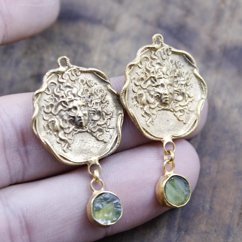 May include: A pair of gold-coloured earrings. Each earring features a detailed Medusa face design with flowing hair. A small, round, green gemstone hangs below each Medusa pendant, adding a pop of colour and contrast to the design.