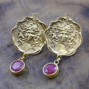 Pode incluir: Um par de brincos dourados. Cada brinco apresenta um design detalhado de rosto com cabelo encaracolado e uma pedra pendurada de cor rubi em forma oval. Os brincos têm uma estética vintage ou antiga.