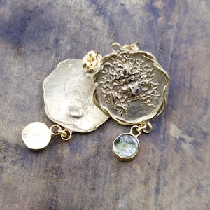 May include: Gold-coloured earrings with a textured, circular design. One earring features a dangling, round, green gemstone. The other earring has a small, round, gold-coloured stud. The earrings are displayed on a wooden surface.