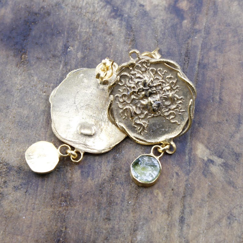 May include: Gold-coloured earrings with a textured, circular design. One earring features a dangling, round, green gemstone. The other earring has a small, round, gold-coloured stud. The earrings are displayed on a wooden surface.