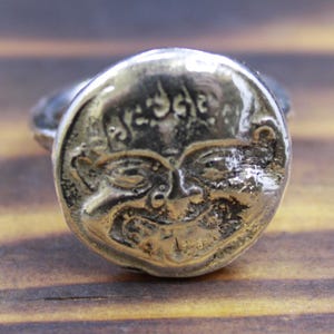 Puede incluir: Anillo plateado con un diseño de cara sonriente. La cara del anillo tiene detalles grabados, incluyendo ojos, nariz y boca sonriente. El anillo está sobre una superficie de madera.