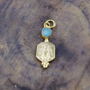 Può includere: Ciondolo color oro con una gemma blu chiaro e un motivo a forma di ape. Il ciondolo ha una forma rettangolare con un'illustrazione dettagliata di un'ape. La gemma è rotonda e si trova sopra il motivo dell'ape. Su una superficie di legno.