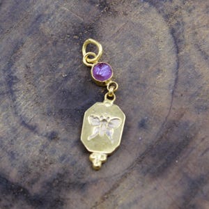 May include: A gold-colored pendant with a butterfly design and a purple gemstone. The pendant has a rectangular shape with a butterfly etched in the center. A small purple gemstone is set above the rectangular shape, connected by a gold chain.