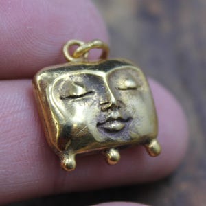 May include: A small, gold-colored pendant with a square face design. The face has closed eyes, a small mouth, and three small ball feet. The pendant has a loop at the top for a chain.