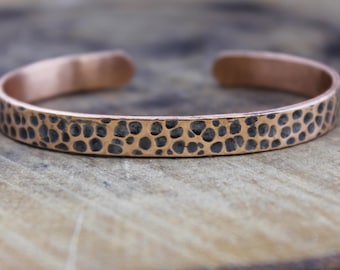 Puede incluir: Una pulsera de puño de cobre con una textura martillada. La pulsera tiene una serie de pequeños puntos oscuros en su superficie. El diseño abierto permite un fácil uso. Esta joya es un accesorio elegante.