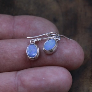 May include: A pair of drop earrings featuring oval-shaped blue opals set in polished silver metal. Each earring has a simple curved hook wire for attachment, showcasing the iridescent light blue and violet flashes within the smooth stone surface.