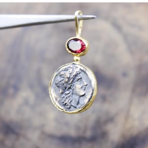 May include: A gold earring with a coin pendant and a red gemstone. The coin has a profile design and a silver finish, set in a gold frame. The oval-shaped gemstone is set in a gold bezel.