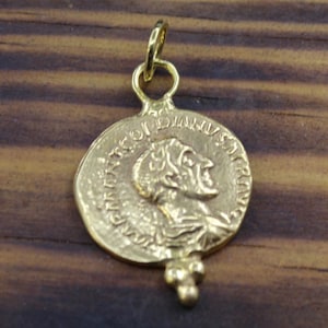 Puede incluir: Colgante dorado con un relieve detallado de una figura histórica en un disco similar a una moneda. El colgante tiene un lazo para una cadena y un elemento decorativo inferior. La superficie de la moneda muestra signos de desgaste, lo que sugiere un estilo antiguo o vintage.