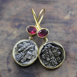 Può includere: Orecchini color oro con pendenti a forma di moneta raffiguranti profili e gemme ovali rosso intenso. Gli orecchini hanno una chiusura a gancio. I pendenti a moneta hanno una finitura color argento e sono incastonati in una montatura color oro.