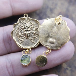 May include: Gold-coloured earrings with a Medusa face design. Each earring has a circular pendant with a detailed face and flowing hair. A small, round, light green stone hangs below one pendant, while the other has a gold-coloured disc.