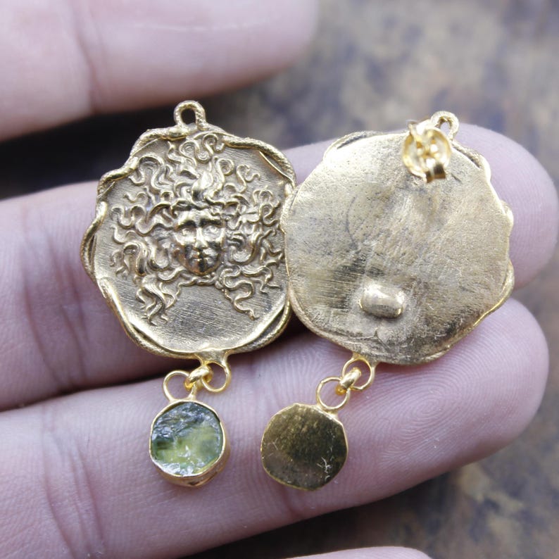 May include: Gold-coloured earrings with a Medusa face design. Each earring has a circular pendant with a detailed face and flowing hair. A small, round, light green stone hangs below one pendant, while the other has a gold-coloured disc.