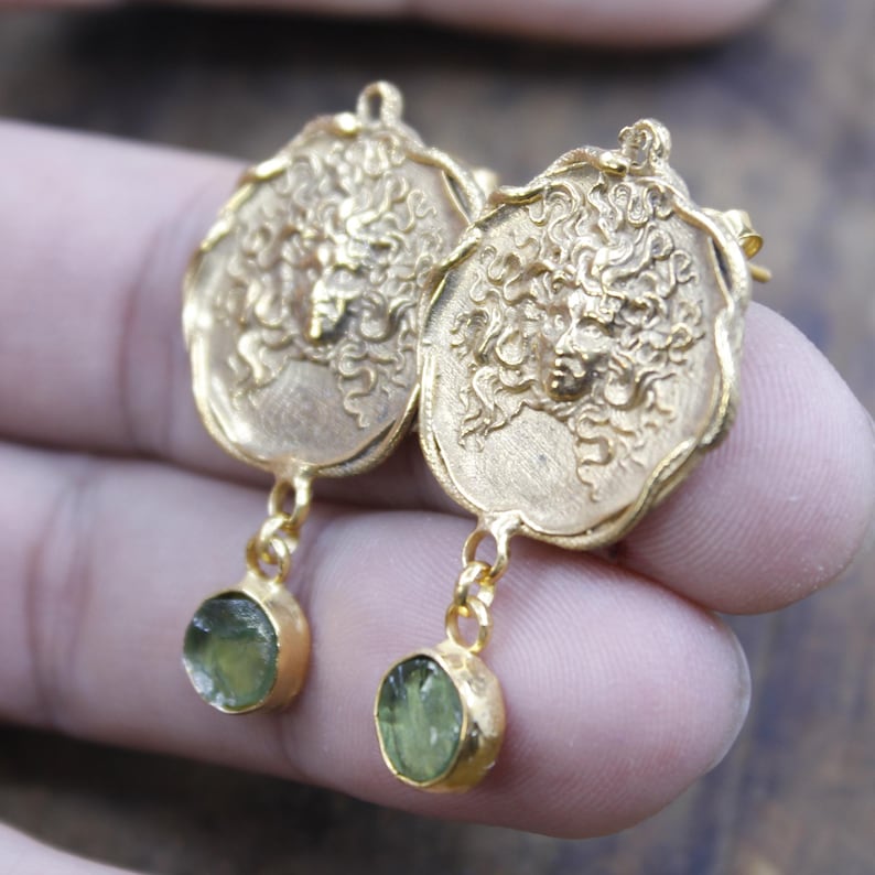 May include: Gold-coloured earrings with an oval pendant featuring a detailed face design. Each earring has a small, round, light green gemstone hanging below. The earrings are held up by a person's fingers.