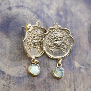 May include: A pair of gold-coloured earrings. Each earring has a detailed, textured design with a circular pendant and a small, round, light green stone dangling below. The earrings are displayed on a textured, neutral-toned surface.