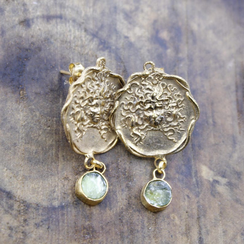 May include: A pair of gold-coloured earrings. Each earring has a detailed, textured design with a circular pendant and a small, round, light green stone dangling below. The earrings are displayed on a textured, neutral-toned surface.
