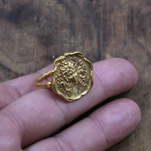May include: A gold-toned ring featuring a circular, scalloped-edge face with a detailed, embossed relief of a face surrounded by flowing, snake-like hair. The band is thin and connects to the side of the textured, coin-shaped centerpiece.