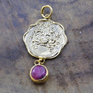 May include: A gold-colored pendant featuring a detailed face design with flowing hair. A round, deep pink gemstone hangs below the pendant. The pendant is displayed on a wooden surface.