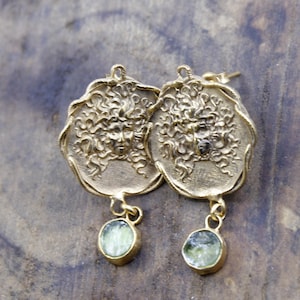 May include: Gold-coloured earrings with a classical face design and flowing hair. Each earring has a circular pendant with a green stone. The earrings are designed with a post-back closure.
