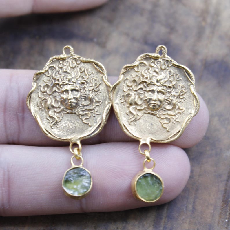 May include: A pair of gold earrings. Each earring has a detailed face design with flowing hair, and a small, oval, green gemstone pendant. The earrings are designed with a loop at the top for hanging.