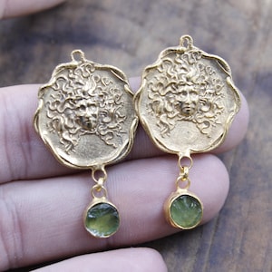 May include: A pair of gold-coloured earrings. Each earring features a detailed face design with flowing hair, and a small, round, green gemstone pendant. The earrings are displayed against a neutral background.