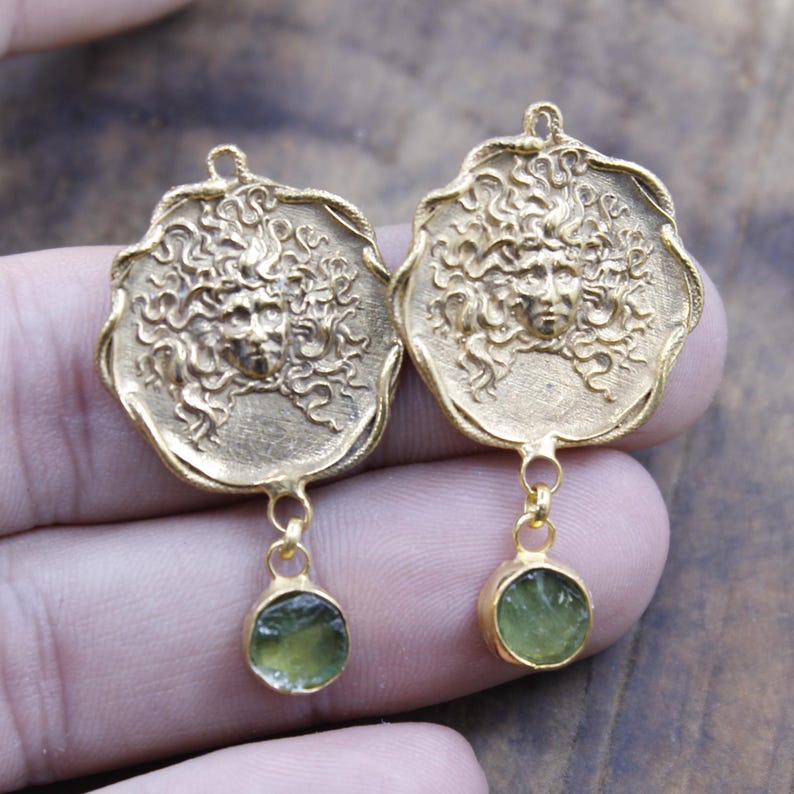May include: A pair of gold-coloured earrings. Each earring features a detailed face design with flowing hair, and a small, round, green gemstone pendant. The earrings are displayed against a neutral background.