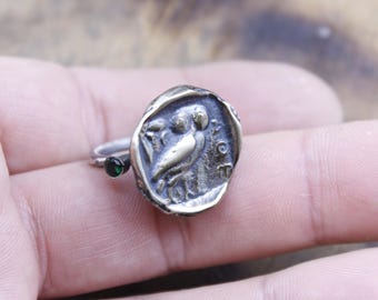 May include: A silver ring featuring a detailed owl design within a circular frame. The ring has a small, round, green gemstone on the band. The owl is depicted in a classical style, with intricate detailing on its feathers and features.