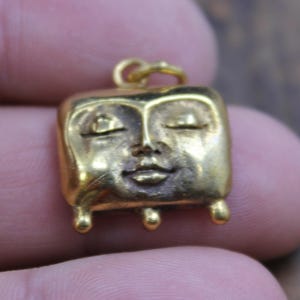 May include: A small, gold-colored pendant with a square face design. The face has closed eyes, a small nose, and a closed mouth. The pendant has a loop at the top for a chain and three small, rounded feet.