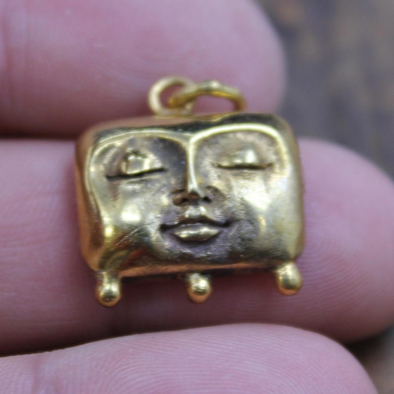 May include: A small, gold-colored pendant with a square face design. The face has closed eyes, a small nose, and a closed mouth. The pendant has a loop at the top for a chain and three small, rounded feet.