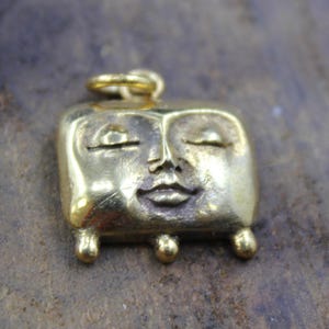 May include: A gold-colored pendant with a smiling face design. The pendant has a rectangular shape with rounded corners, closed eyes, and small, ball-shaped feet. A small loop is at the top for a chain.