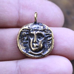 Puede incluir: Colgante redondo de bronce con un relieve detallado de un rostro, posiblemente una figura clásica. El colgante tiene un lazo en la parte superior para una cadena. El metal tiene un aspecto envejecido y antiguo.