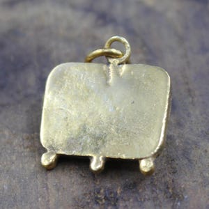May include: A gold-colored pendant with a rectangular body and three small, rounded feet. A loop is attached at the top for a chain. The pendant has a slightly aged appearance.