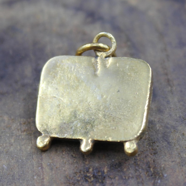 May include: A gold-colored pendant with a rectangular body and three small, rounded feet. A loop is attached at the top for a chain. The pendant has a slightly aged appearance.