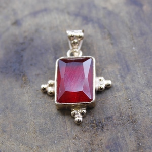 May include: A gold-toned pendant featuring a rectangular, deep red gemstone. The pendant has a decorative design with small, round accents on each side of the gemstone. The setting is a warm, metallic color.