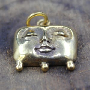 May include: A gold-colored pendant with a smiling face design. The pendant has a rectangular shape with rounded edges and small, ball-shaped feet. The face features closed eyes and a small, smiling mouth. A loop is at the top for hanging.